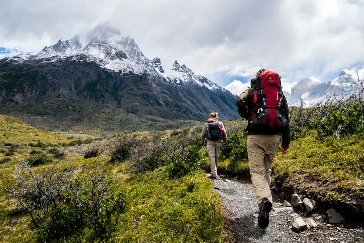 Two hikers walking a mountain trail toward snow covered peaks